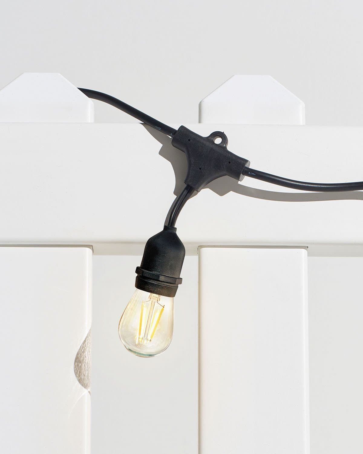 Brightech Ambience Pro outdoor string lights with a lit S14 Edison filament bulb hanging from a black weatherproof socket against a white fence
